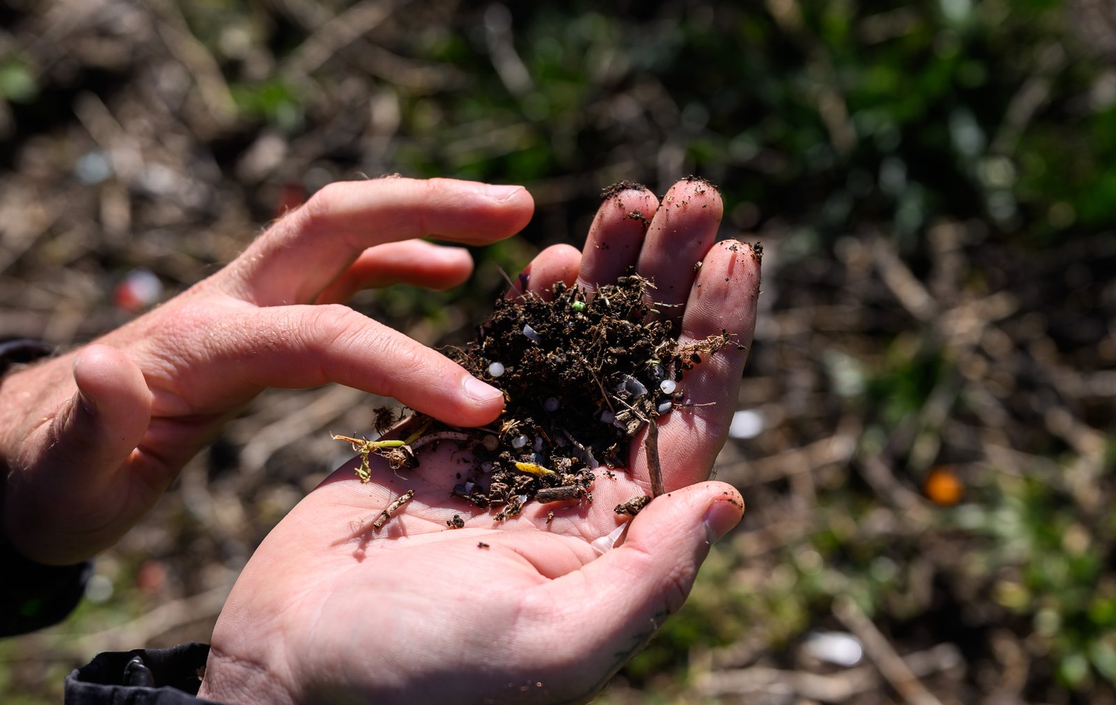 A handful of soil and shoreline debris shot through with fragments of broken plastic